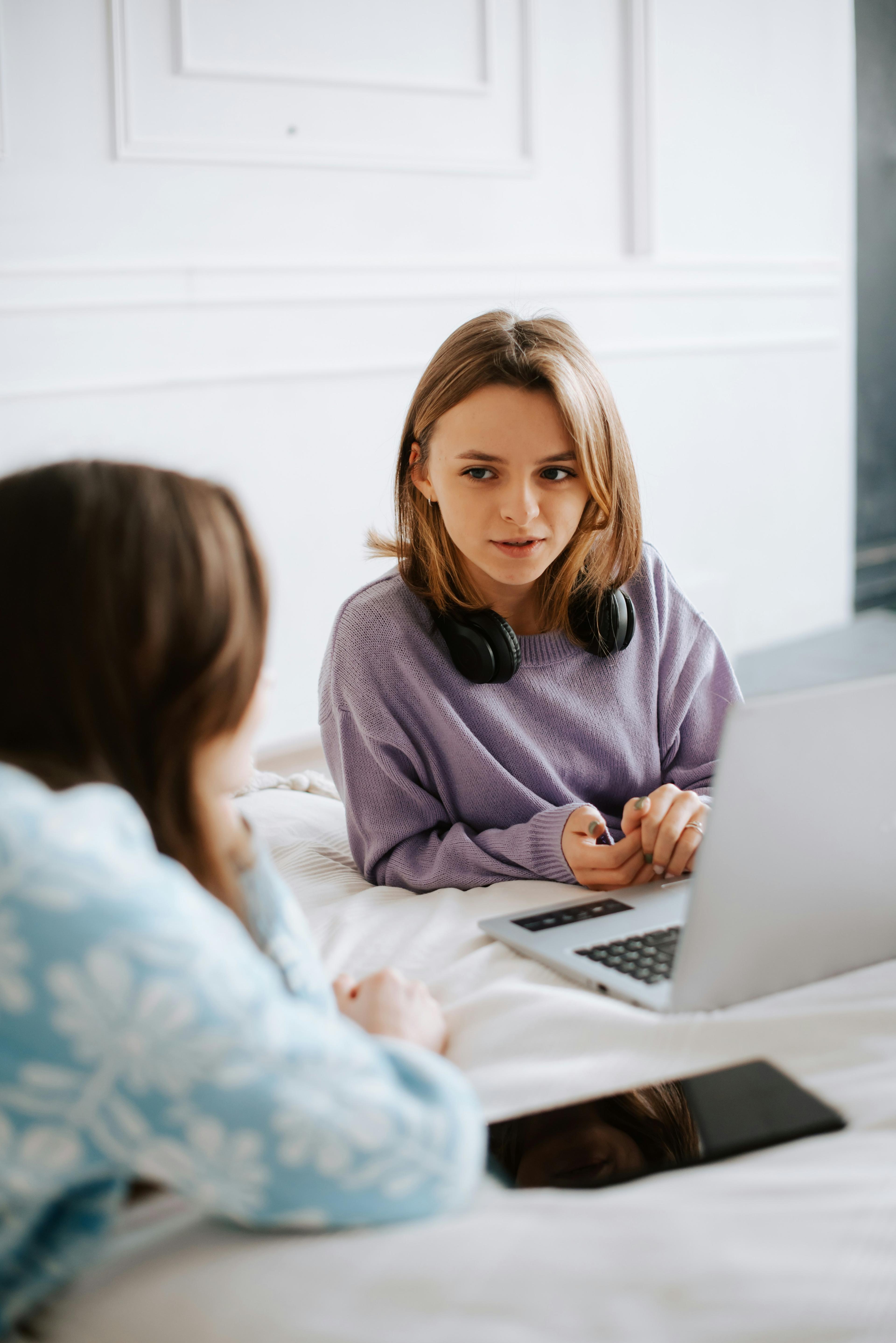 Two young people on a bed in conversation, one with a laptop and headphones round her neck.