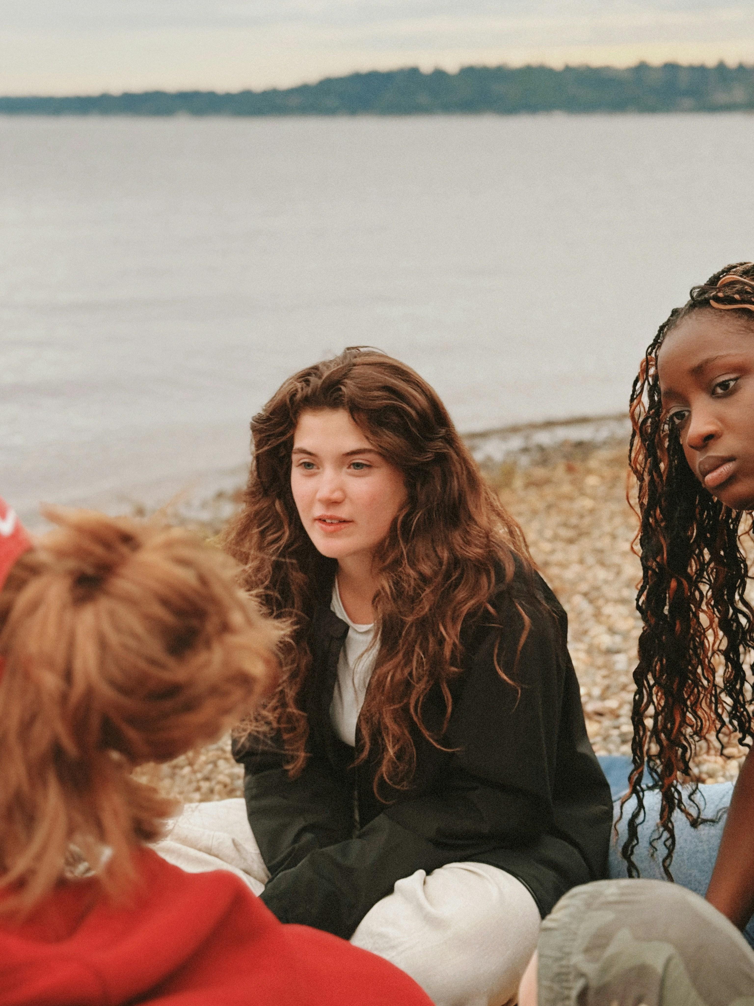 Three young people sitting in a quiet circle on a stony shore, mid-conversation by the water.